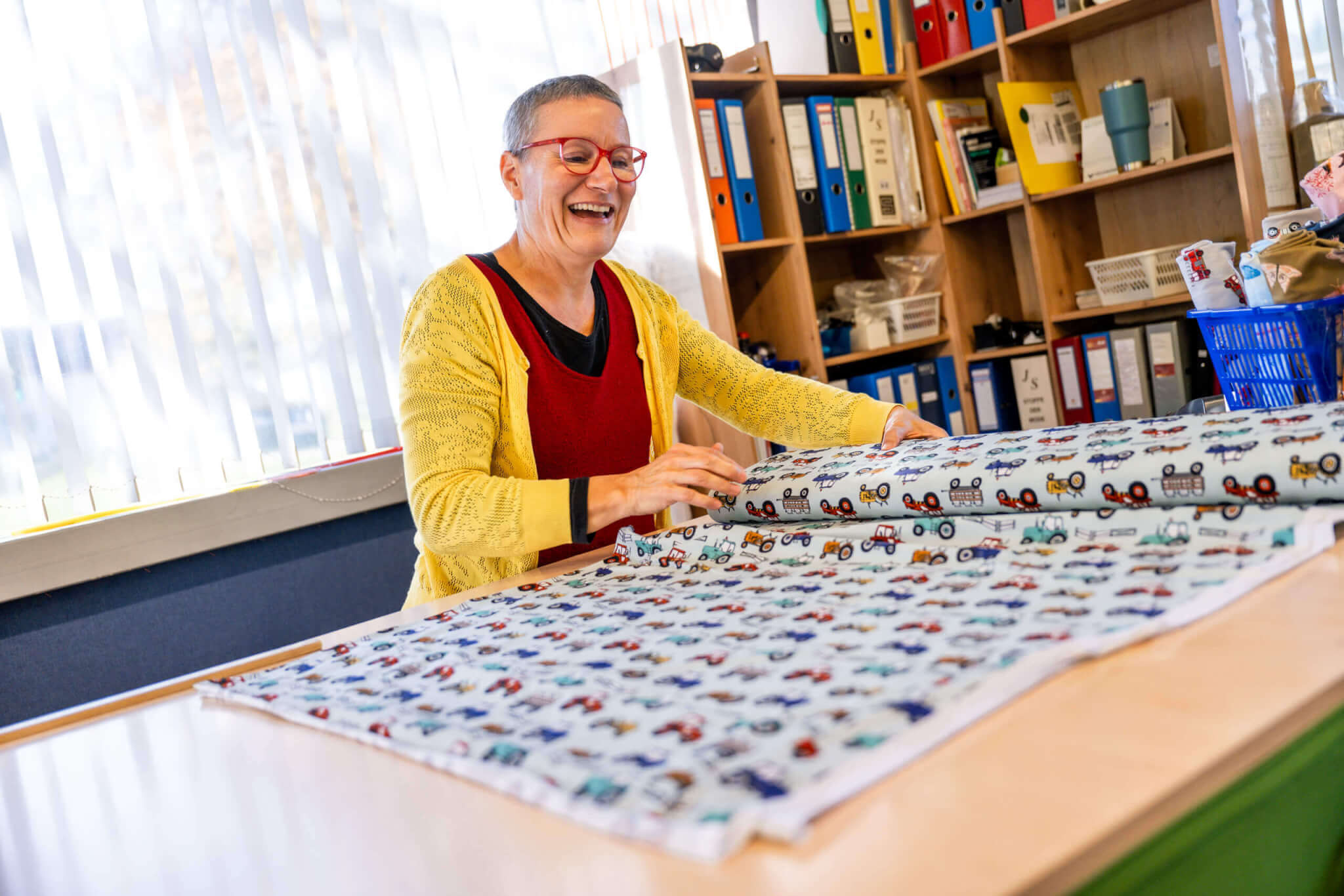 Person mit gelber Strickjacke lacht und breitet Stoff mit buntem Traktormuster auf einem Tisch aus.