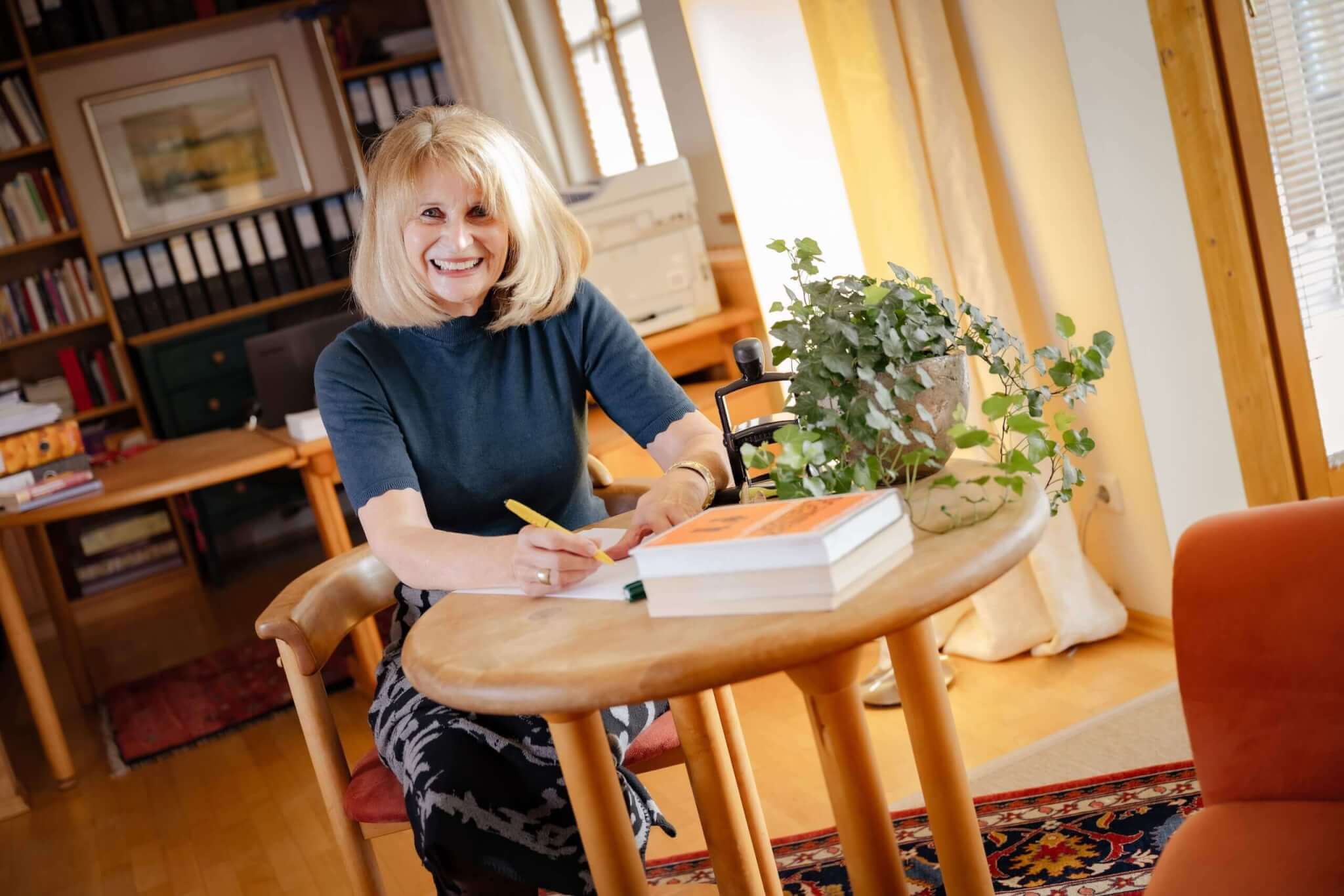 Frau schreibt lächelnd an einem Tisch mit Büchern und Pflanzen in einem gemütlichen, lichtdurchfluteten Raum.