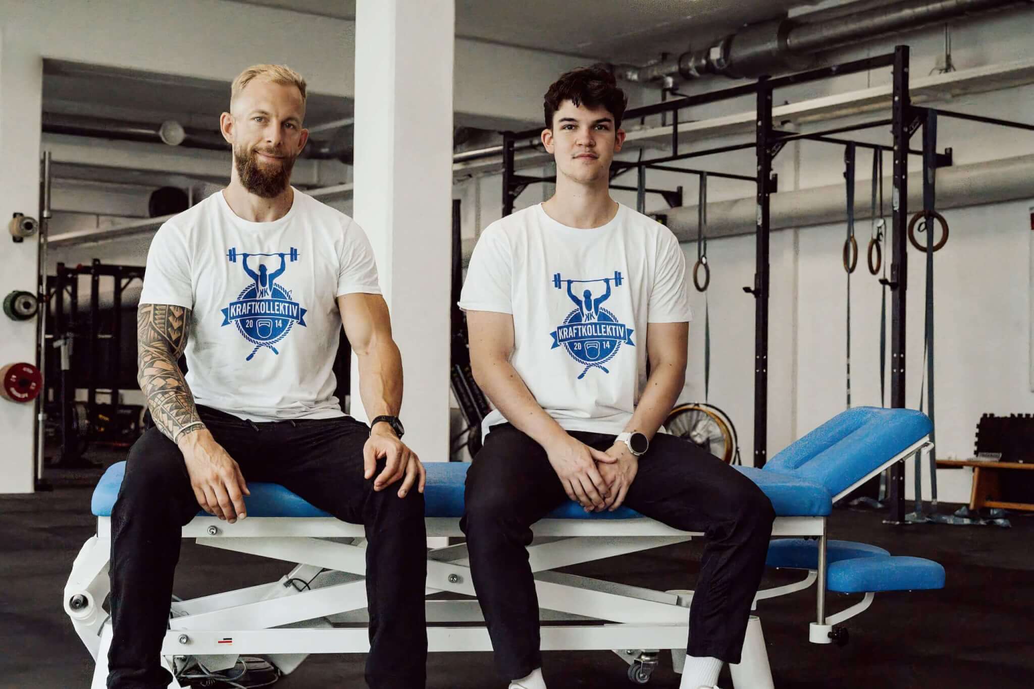 Zwei Männer in einer Turnhalle tragen T-Shirts mit dem Logo Kraftkollektiv und sitzen auf einer Trainingsbank.