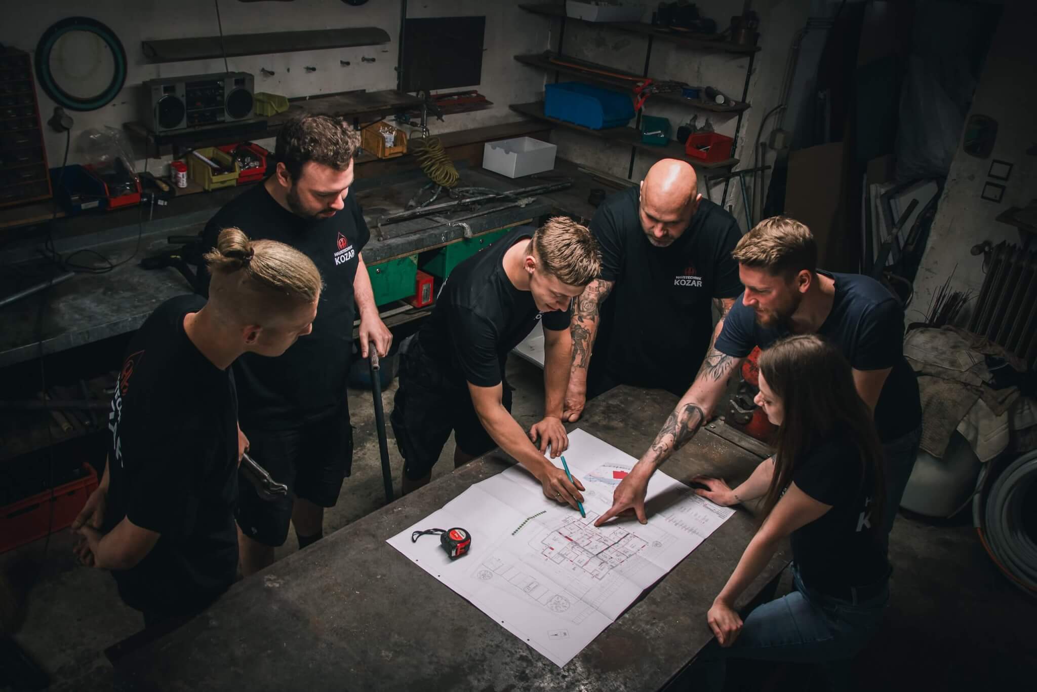 Gruppe von fünf Personen bespricht Baupläne in einer Werkstatt, nahbar und kreativ im Team.